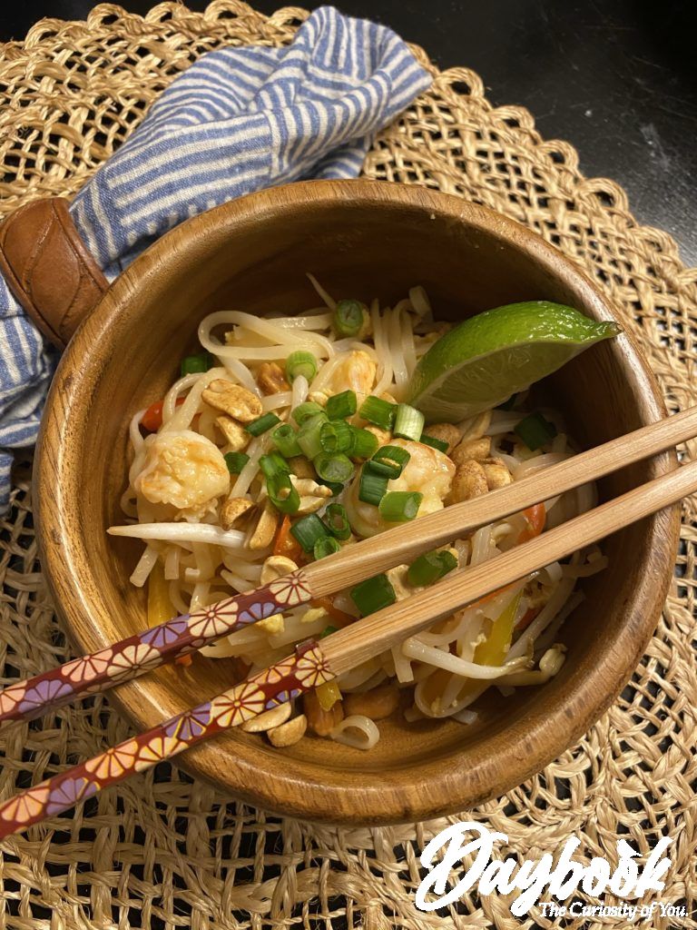 Pad Thai in a wooden bowl with lime on the side and chop sticks across the top.
