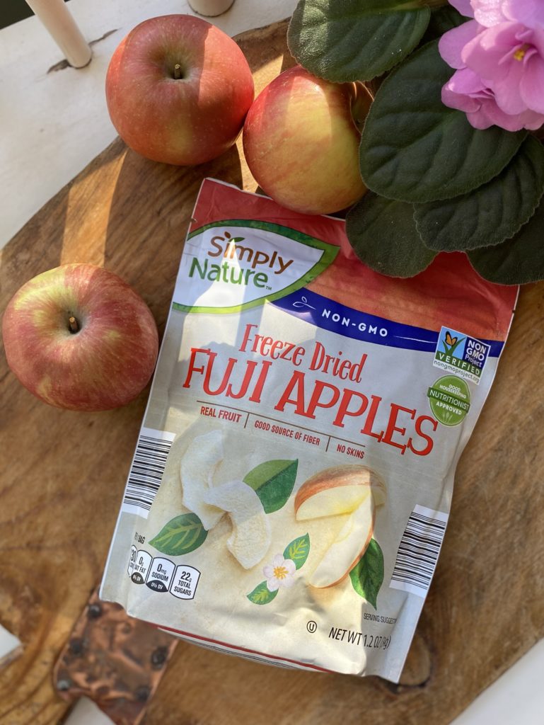 A bag of freeze dried Fuji apples on a cutting board with 3 fresh apples.