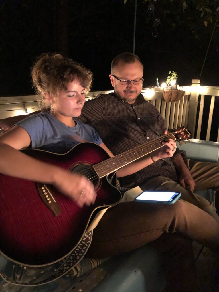 Young girl playing her guitar at night with her dad seated beside her