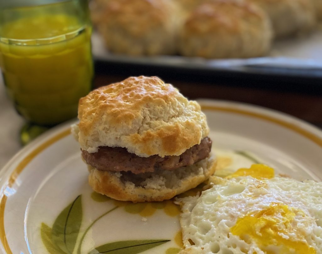 A sausage biscuit with a fried egg on a floral plate next to a glass of orange juice and a pan of warm biscuits.