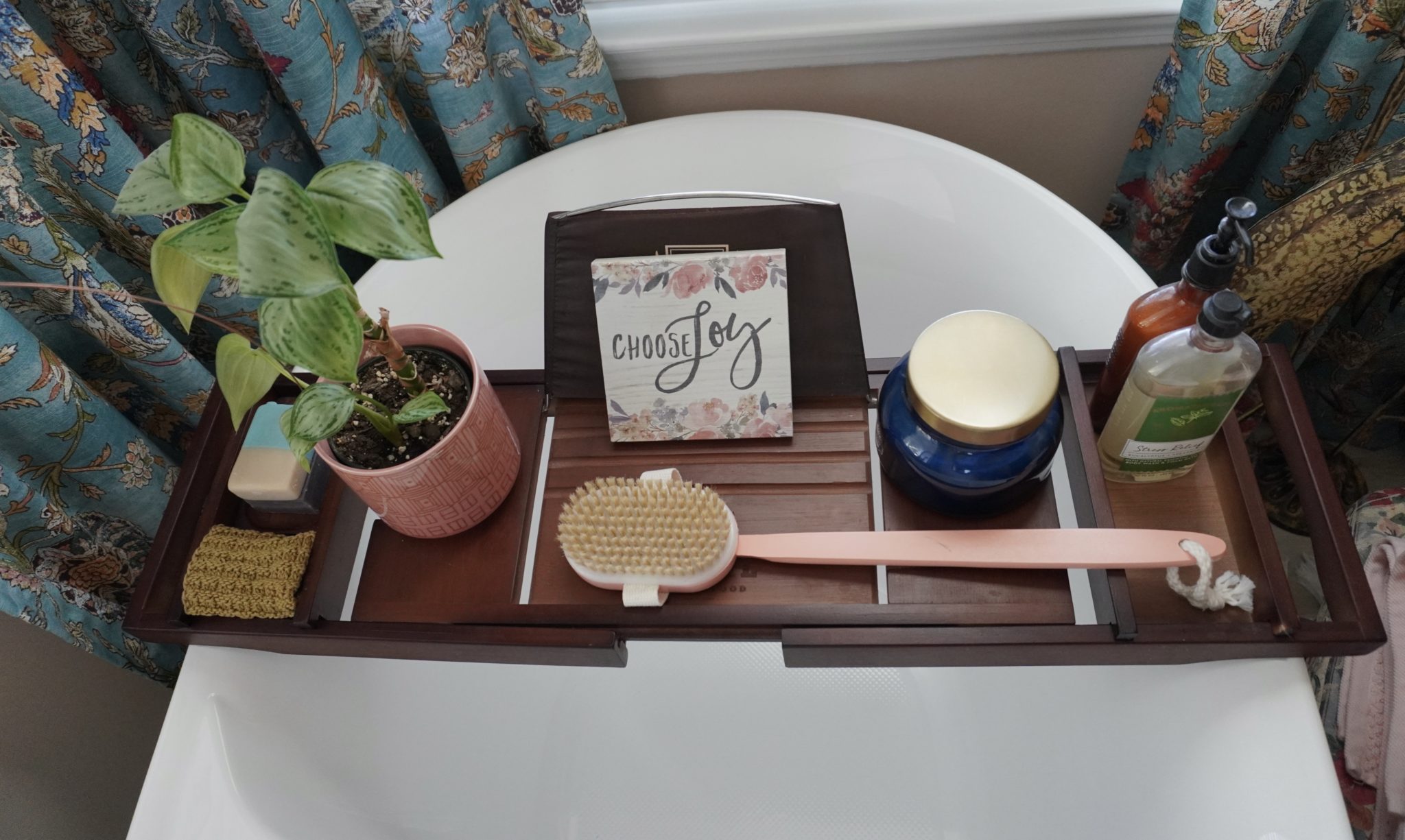 A wooden bath tray over a white soaking tub with a plant and candle on top.