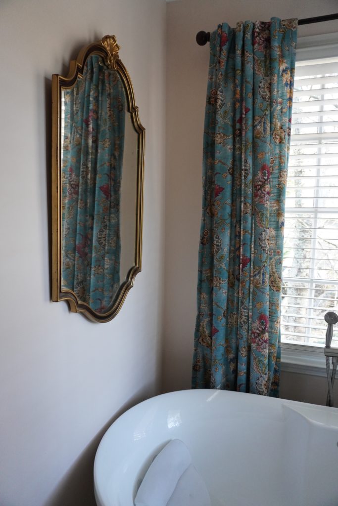 A decorative mirror hanging over a bathtub next to a window.