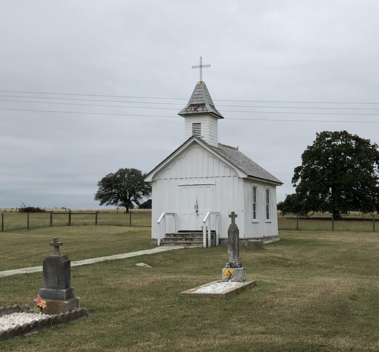 A small white local church in Roud Top Texas with a gravesite in front of the steps.