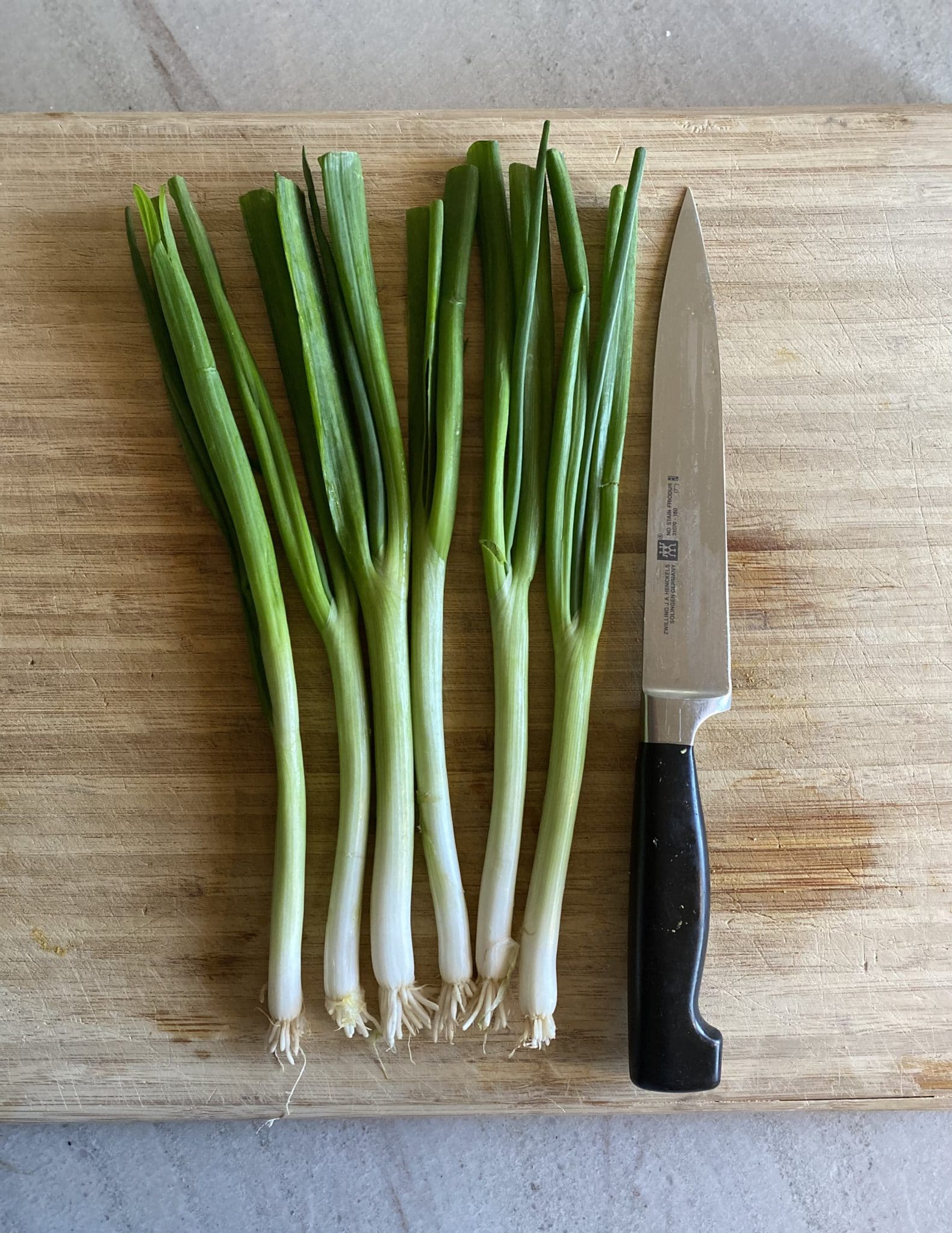 Six green scallions lined up on a wooden cutting board with a chopping knife to the right.