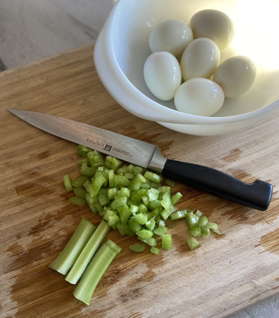 A stalk of celery chopped on a wooden cutting board next to a bowl of boiled eggs.
