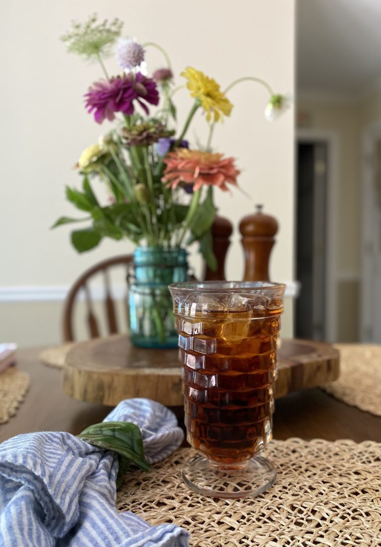 A glass of iced tea on a kitchen table in front of a colorful flower arrangement.