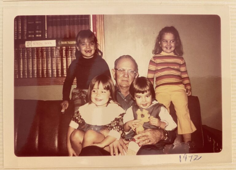 A Pappaw sitting on his brown sofa with his four grandchildren in his lap.