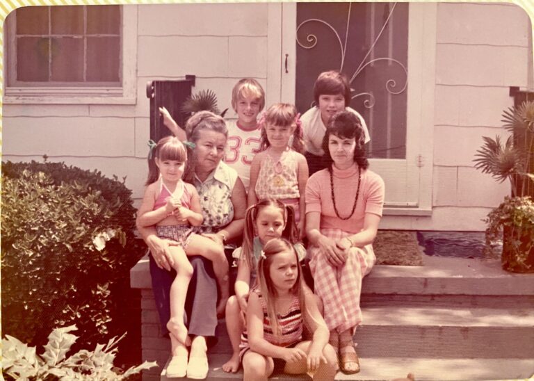 A mother and a grandmother sitting on front porch steps surrounded by six grandchildren.