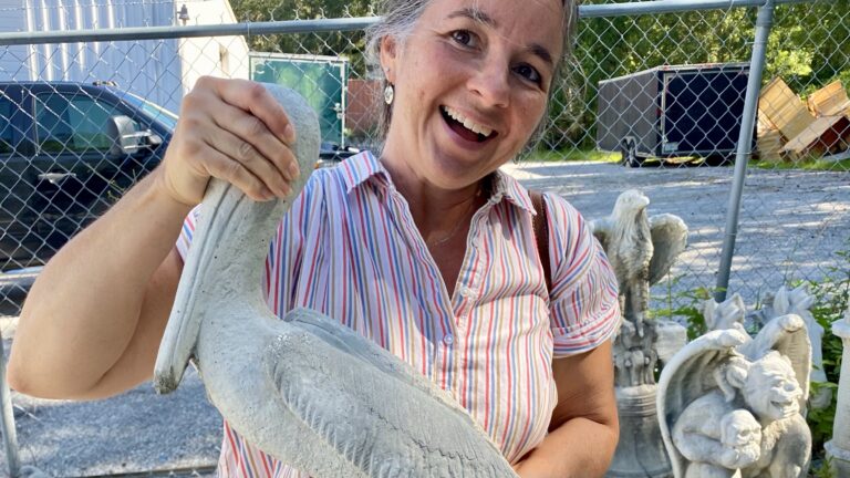 Lady holding a concrete pelican in a parking lot behind a store.