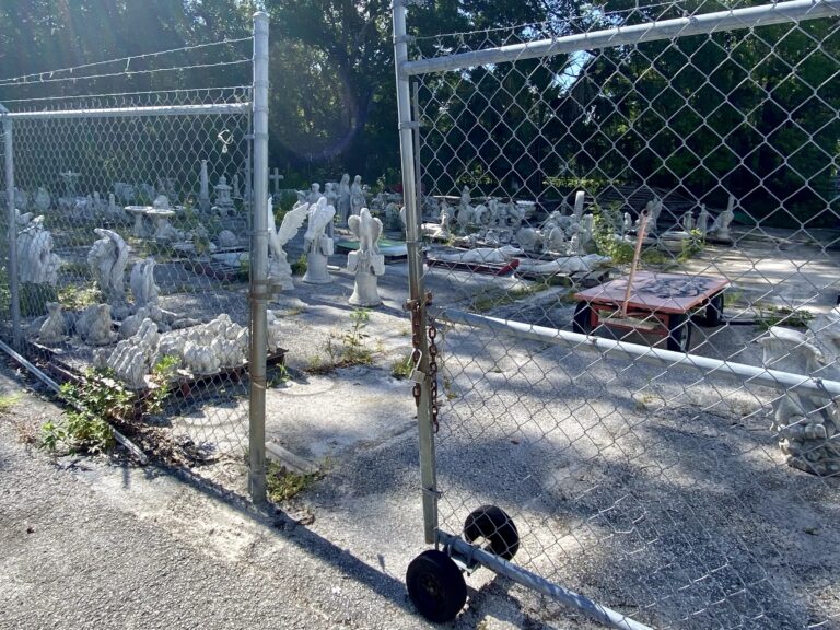 A parking lot with pallets of concrete pelicans and other figures behind a chain-linked fence.