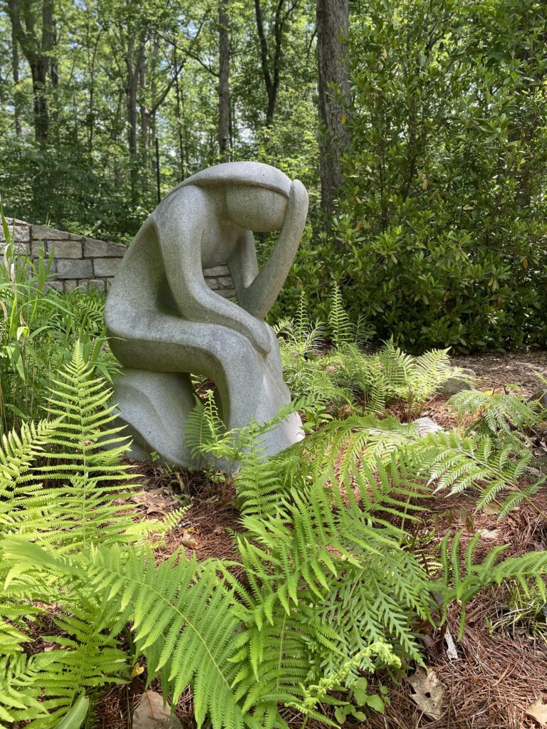 A large concrete statue of a lady sitting and holding her head down in a garden of ferns.