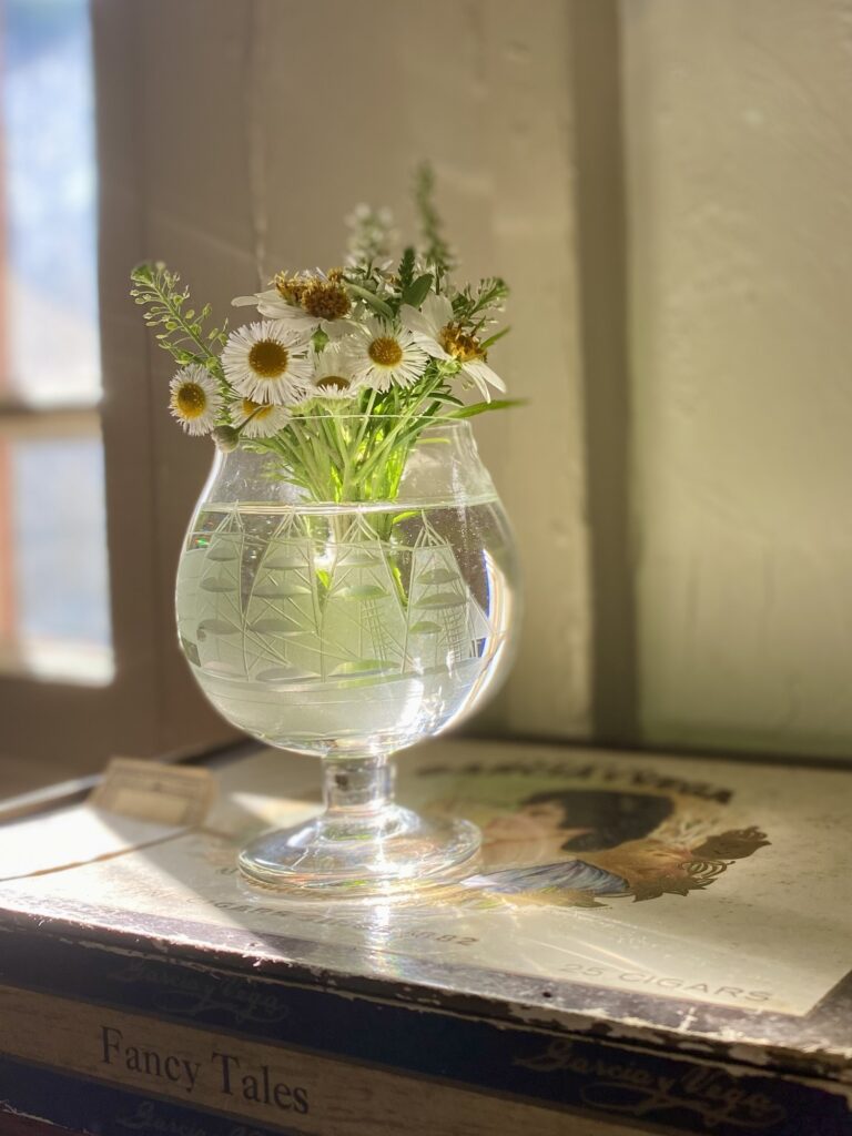 A bundle of wildflowers in a tiny bouquet placed in a glass stemmed whiskey glass that has a ship etched on the glass.